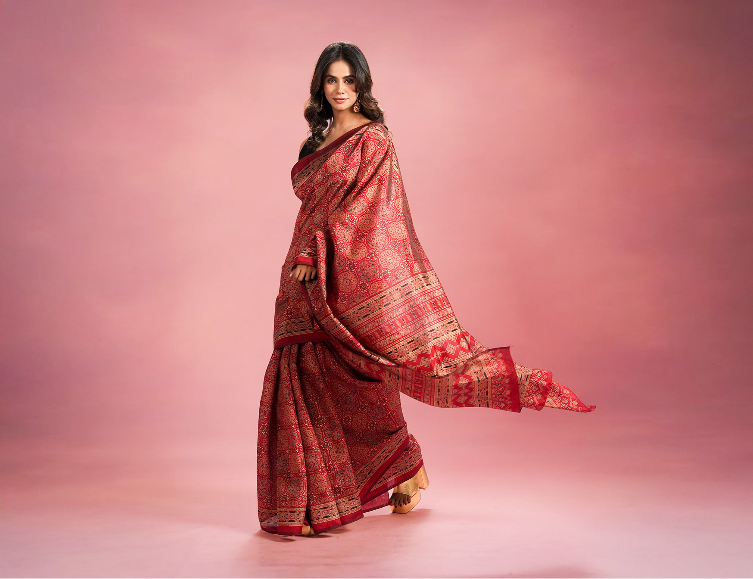 Woman in a traditional red saree with intricate ethnic patterns, posing gracefully against a soft pink gradient background